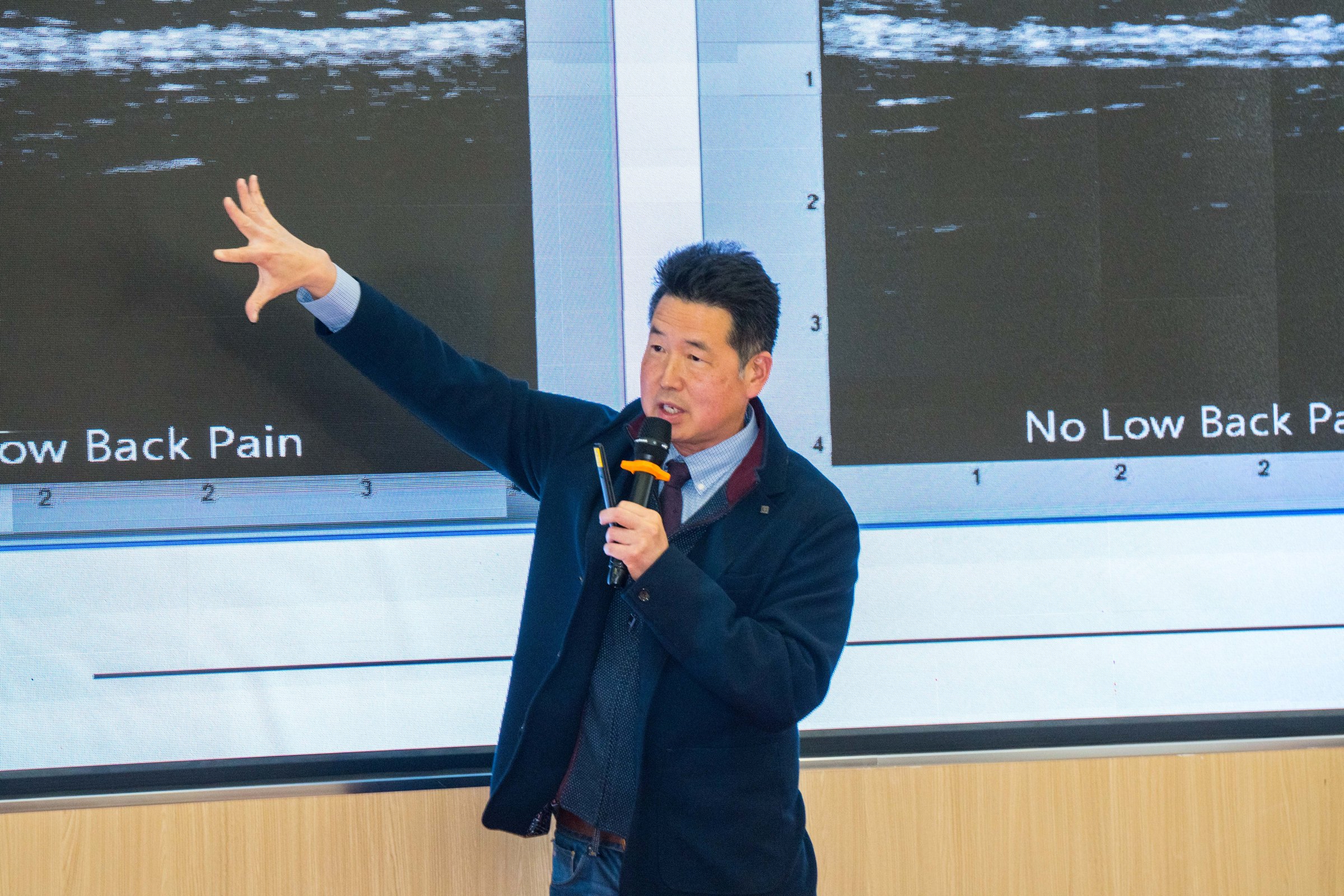 Dr. Koh holding a microphone and gesturing toward ultrasound images on a large screen during a lecture on low back pain